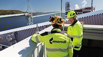 two men in hi-viz clothing overlook a production site with a river and forest in the background