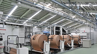 Industrial production line with large rolls of brown paper mounted on automated unwinding machines inside a factory hall.