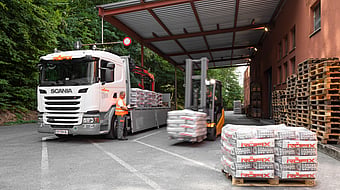 Delivery truck parked under a covered loading area while a forklift transports wrapped pallets. Stacks of palletised building materials are arranged in the foreground, with additional wooden pallets stored beside an industrial building surrounded by trees.