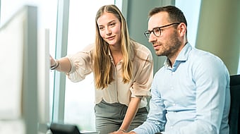 A man and a woman look closely at a computer screen; the woman points at it while both appear highly focused and engaged.