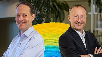 Two people standing with folded arms in front of a colourful circular display, with indoor plants and office elements visible in the background.